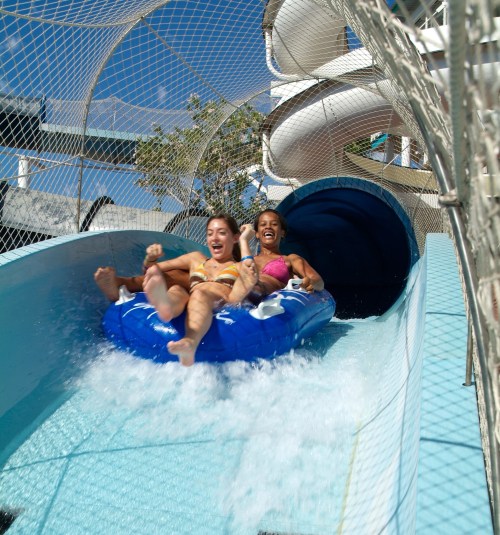 Two women joyfully slide down a water slide on a blue inflatable raft, splashing into the water below.