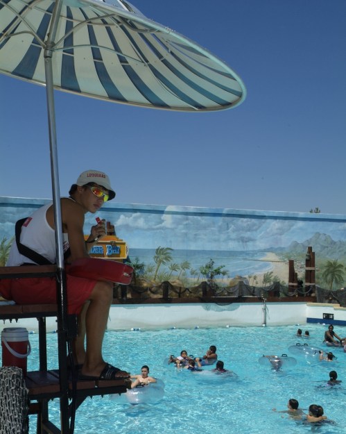 A lifeguard sits on a tall chair by a pool, watching swimmers below under a striped umbrella on a sunny day.