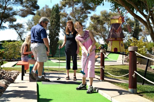 A family enjoys mini-golf on a sunny day, with a colorful windmill in the background and lush greenery around.