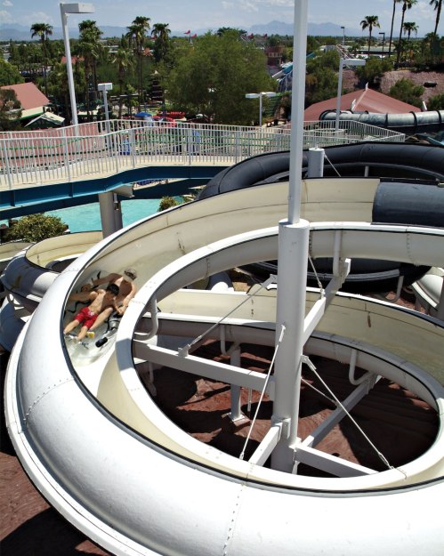 Two people slide down a water slide in a theme park, surrounded by palm trees and other attractions.