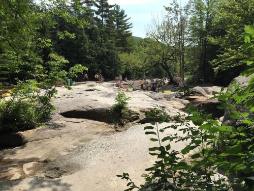 A rocky area by a river surrounded by trees, with people enjoying the outdoors on a sunny day.
