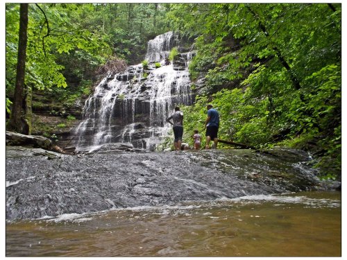 Three people stand near a waterfall surrounded by lush green trees, with water cascading down rocky terrain.