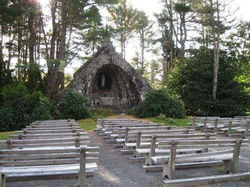 A stone chapel with a statue, surrounded by trees and wooden benches arranged for seating.
