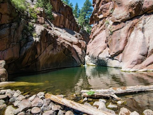 A serene natural landscape featuring a calm pool surrounded by rocky cliffs and trees under a clear blue sky.