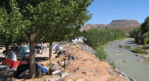 A scenic campsite by a river, featuring tents, trees, and rocky banks with mountains in the background under a clear blue sky.