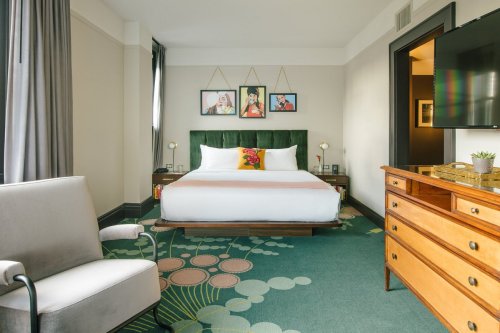 A stylish hotel room featuring a green bed, modern decor, artwork on the walls, and a cozy seating area.