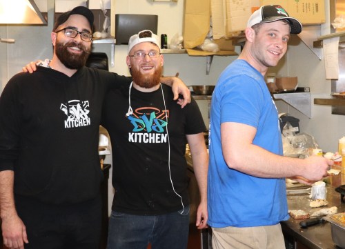 Three men smiling in a kitchen, wearing matching shirts, engaged in food preparation.