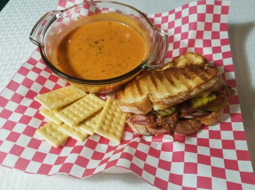 A bowl of tomato soup, a grilled sandwich with pickles, and crackers on a checkered paper plate.