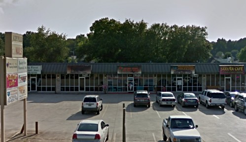 A small shopping plaza with several storefronts and parked cars in front, surrounded by trees.