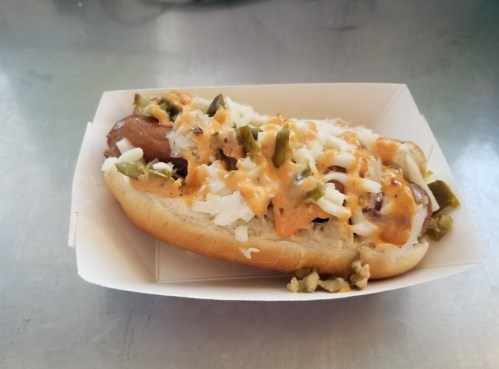 A hot dog in a bun topped with cheese, onions, and jalapeños, served in a paper tray on a metal surface.