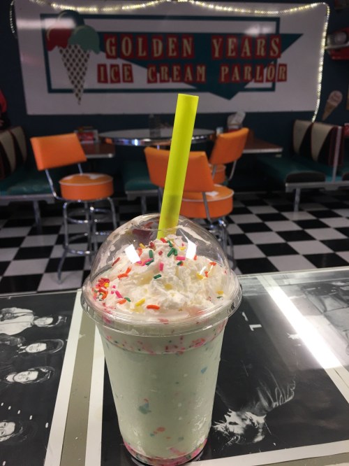 A colorful milkshake topped with whipped cream and sprinkles, set in a retro ice cream parlor with orange seating.