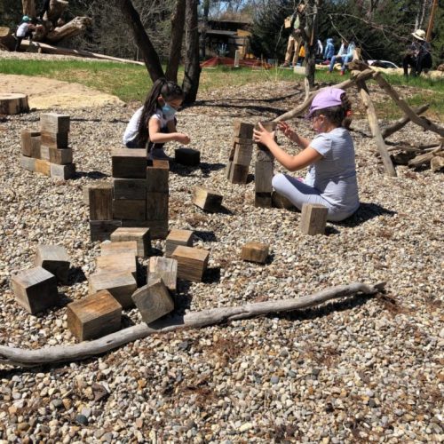 Two children build with wooden blocks on a gravel surface, surrounded by nature and other people in the background.