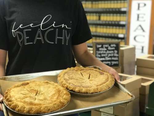 A person in a "feelin' peachy" shirt holds a tray with two freshly baked peach pies.