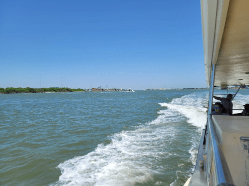 A view from a boat showing a calm waterway with a wake, under a clear blue sky. Passengers are visible on the side.