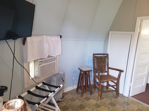 Cozy room with a TV, air conditioner, wooden chair, and a small stool, featuring light-colored walls and a closet.