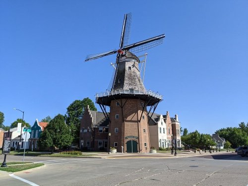 A tall windmill stands prominently against a clear blue sky, surrounded by green trees and charming buildings.