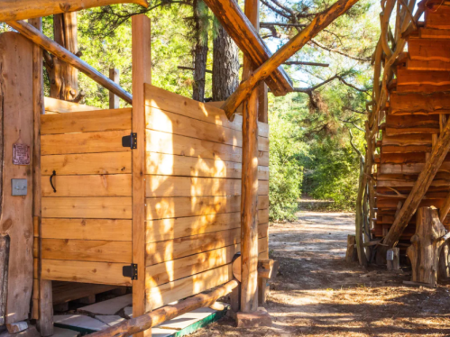Wooden structure with a rustic design, featuring a staircase and an enclosed area, surrounded by trees.
