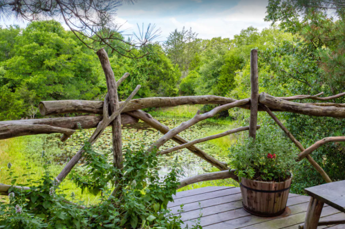 A wooden railing overlooks a lush green landscape with a pond and trees, featuring a potted plant in the foreground.