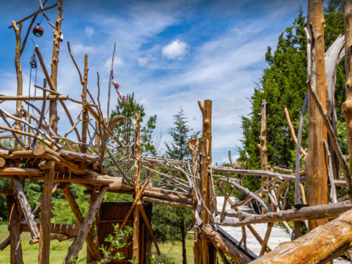 A whimsical wooden structure with interconnected pathways and trees under a blue sky with scattered clouds.