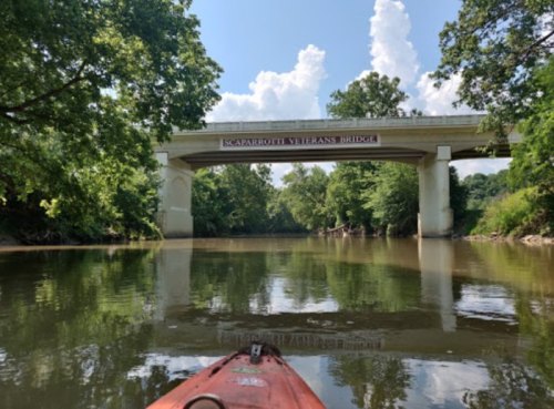 A view from a kayak on a river, with a bridge overhead surrounded by lush greenery and blue skies.