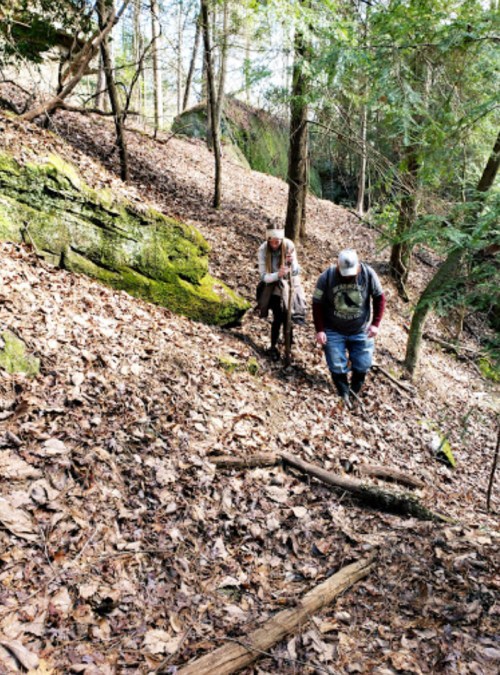 Two hikers navigate a leaf-covered trail in a wooded area, surrounded by trees and rocky terrain.