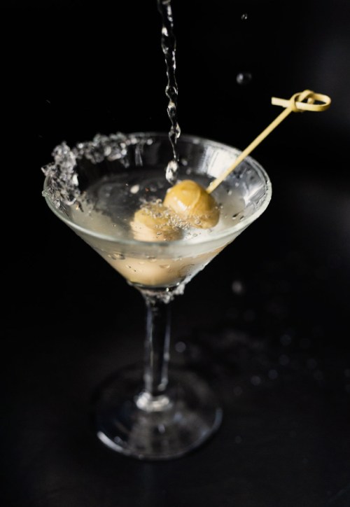 A martini glass with olives, water splashing into it against a dark background.