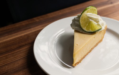 A slice of lime cheesecake topped with whipped cream and a lime wedge on a white plate, resting on a wooden table.
