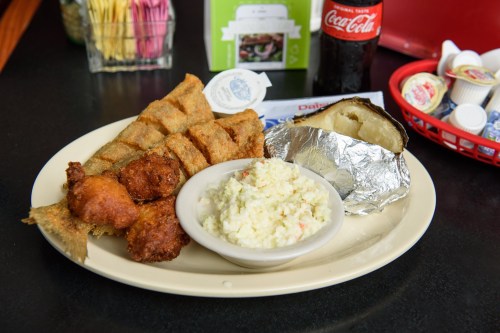 A plate of fried fish, hushpuppies, coleslaw, and a baked potato, with a soda in the background.