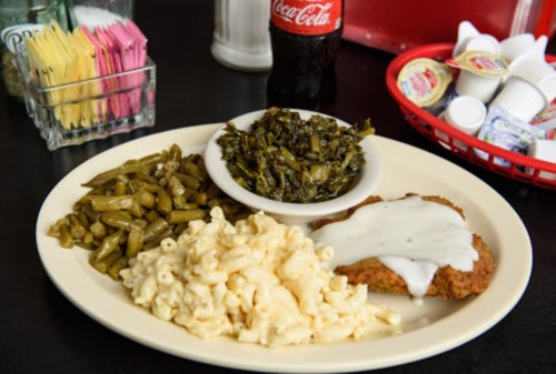 A plate with fried chicken topped with gravy, macaroni and cheese, green beans, and collard greens, with a soda in the background.