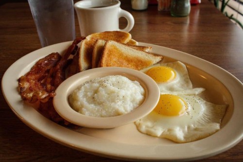 A breakfast plate with two eggs, grits, bacon, and toast, accompanied by a cup of coffee and a glass of water.