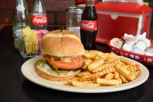 A plate with a burger topped with lettuce and tomato, served with crinkle-cut fries and drinks in the background.