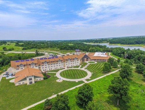 Aerial view of a large hotel complex surrounded by green fields and a lake, featuring landscaped grounds and pathways.