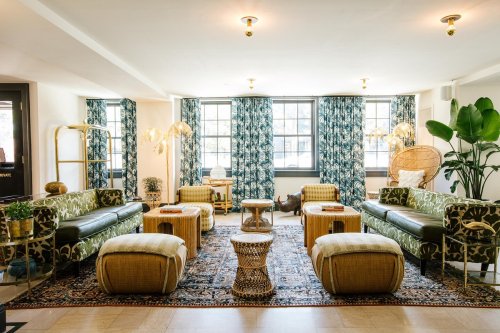 A stylish lounge with patterned curtains, green sofas, woven stools, and decorative plants, creating a cozy atmosphere.