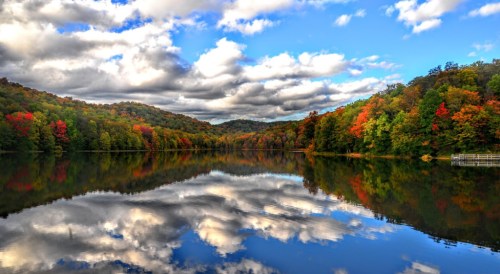 A serene lake surrounded by vibrant autumn foliage and fluffy clouds reflecting on the water's surface.