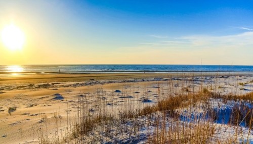A serene beach scene at sunset, with gentle waves, sandy shore, and tall grass in the foreground.