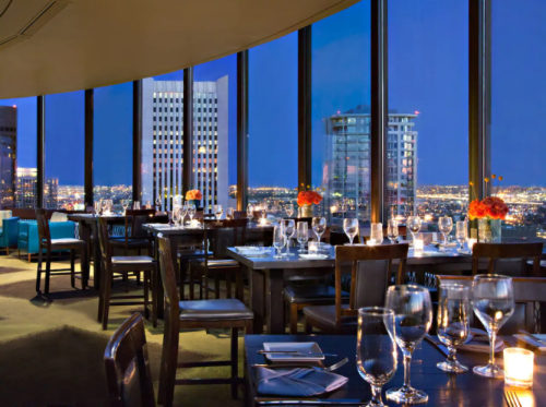 Elegant restaurant interior with city skyline views at dusk, featuring tables set with glassware and floral centerpieces.