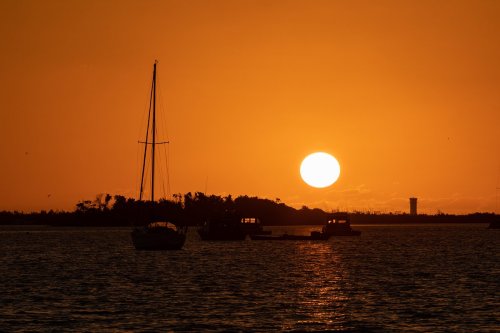 Silhouette of boats and a sailboat against a vibrant orange sunset over calm waters.
