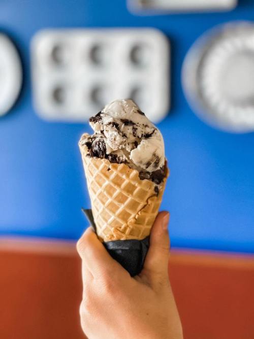 A hand holds a waffle cone filled with chocolate and cookies and cream ice cream against a blue wall.