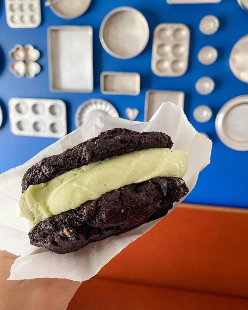 A hand holding a chocolate cookie sandwich filled with green ice cream, against a blue background with various baking trays.