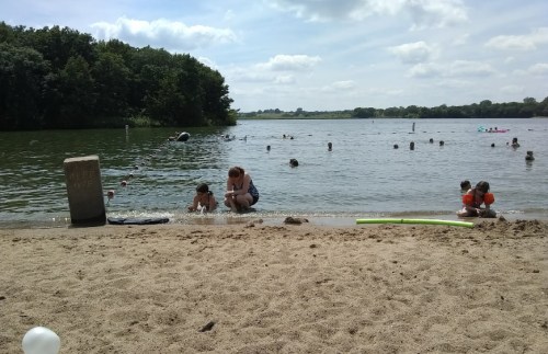 A sandy beach by a lake with children playing in the water and adults supervising under a sunny sky.