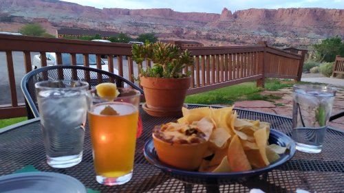 A table with a beer, two glasses of water, and a bowl of chili with tortilla chips, set against a scenic mountain backdrop.