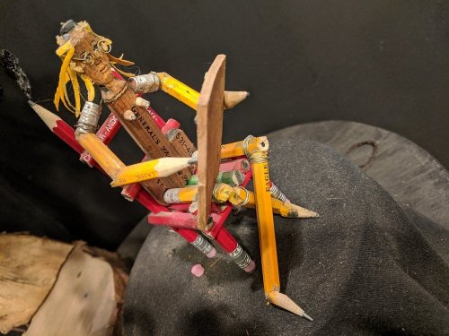 A creative sculpture made from colored pencils, resembling a figure sitting on a rock.