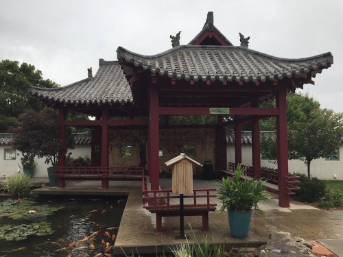 A traditional Chinese pavilion with a tiled roof, surrounded by a pond filled with koi fish and lush greenery.