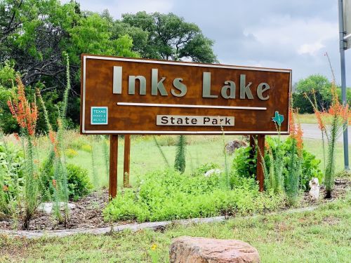 Sign for Inks Lake State Park, featuring green grass and colorful plants, under a cloudy sky.