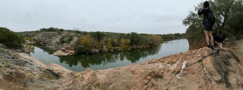 A person stands on a rocky ledge by a river, with a dog nearby, surrounded by trees and autumn foliage.