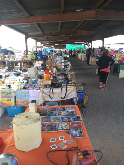 A bustling market scene with various vendor tables displaying crafts, collectibles, and people browsing in the background.