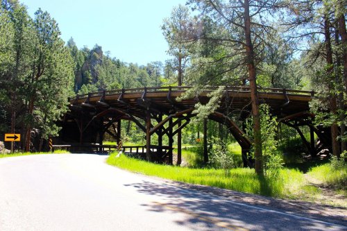 A wooden bridge curves over a road, surrounded by lush green trees and rocky hills under a clear blue sky.