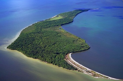 Aerial view of a lush, green island curving into a blue ocean, with sandy shores and varying water colors.