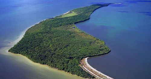 Aerial view of a lush, green island extending into a blue body of water, with a sandy shoreline visible.