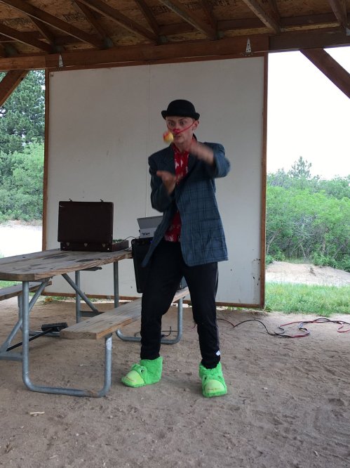 A performer in a bowler hat and colorful outfit juggles while standing on a stage with a picnic table in the background.